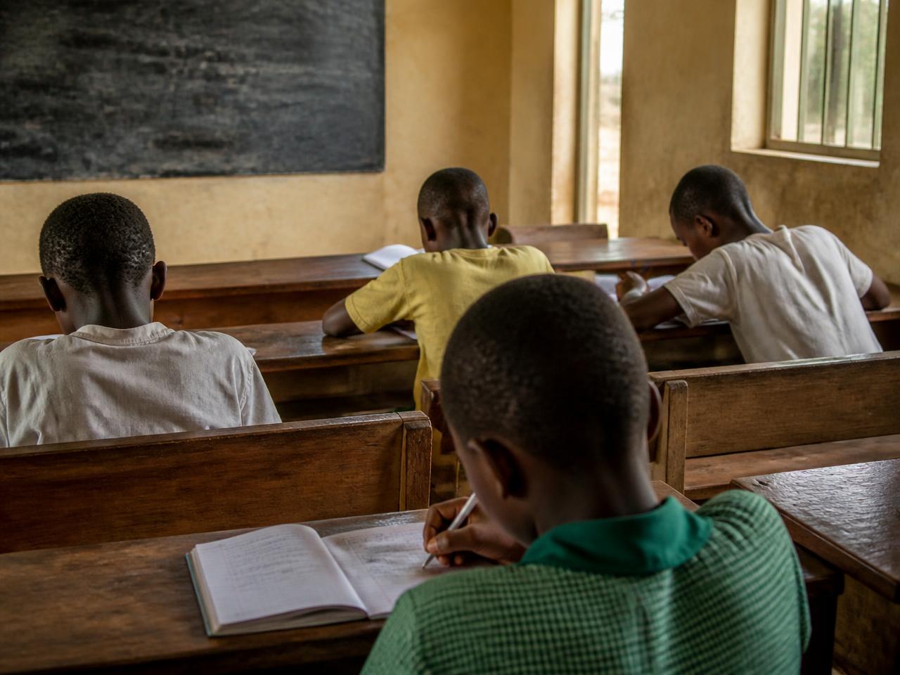 Children seen from behind writing in exercise books in a simple sunlit classroom